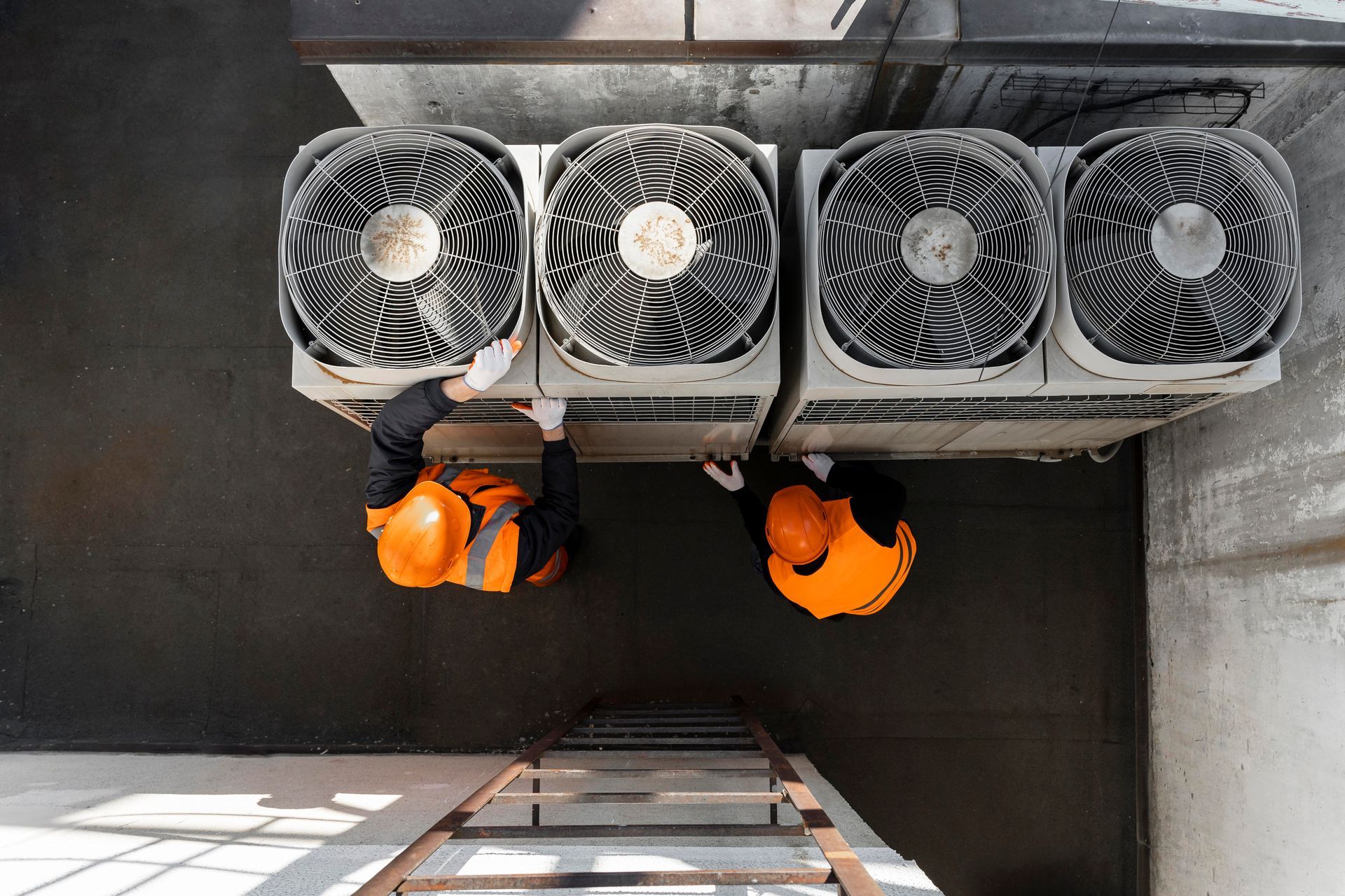 Two people in orange vests and hardhats work on rooftop air conditioning units.