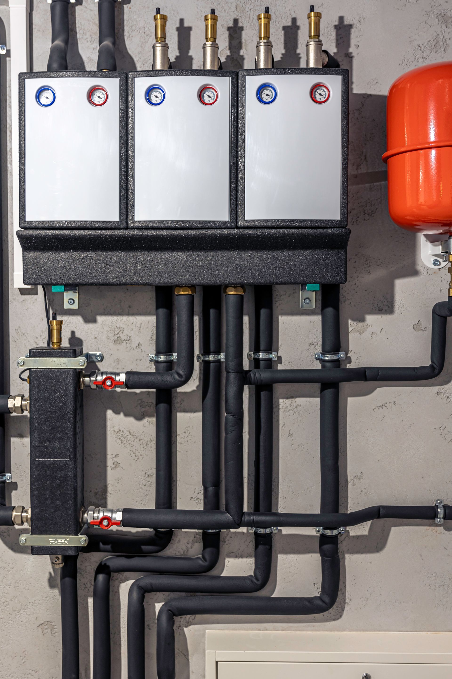 Plumbing system with insulated pipes, silver panels, and an orange expansion tank mounted on a concrete wall.