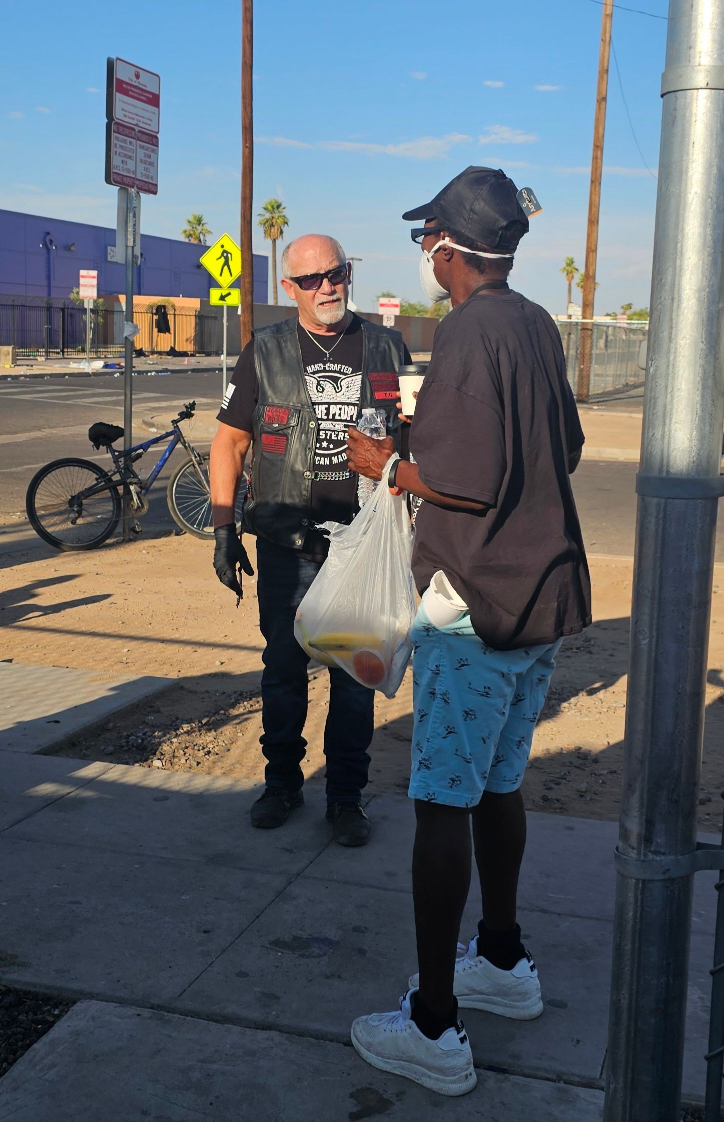 A man in a vest hands a bag to a person on a sidewalk near a road and bicycle.