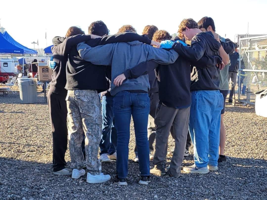 A group of young people with arms around each other in a circle outdoors, a sign of unity.