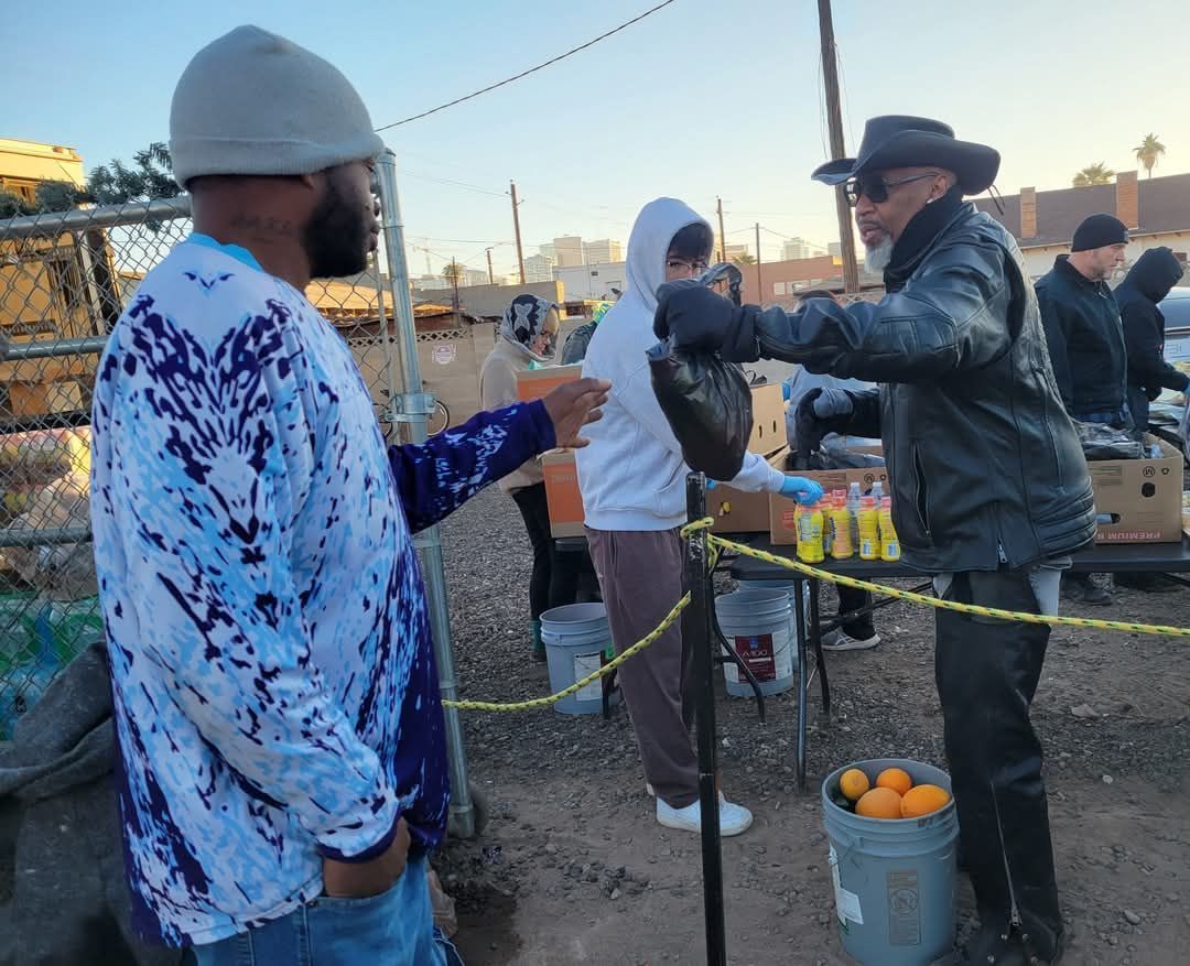 People serving food outdoors. Man in hat hands a drink to another.