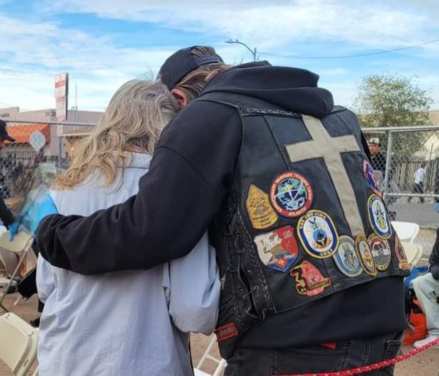 A man wearing a leather vest with a cross on it is hugging a woman