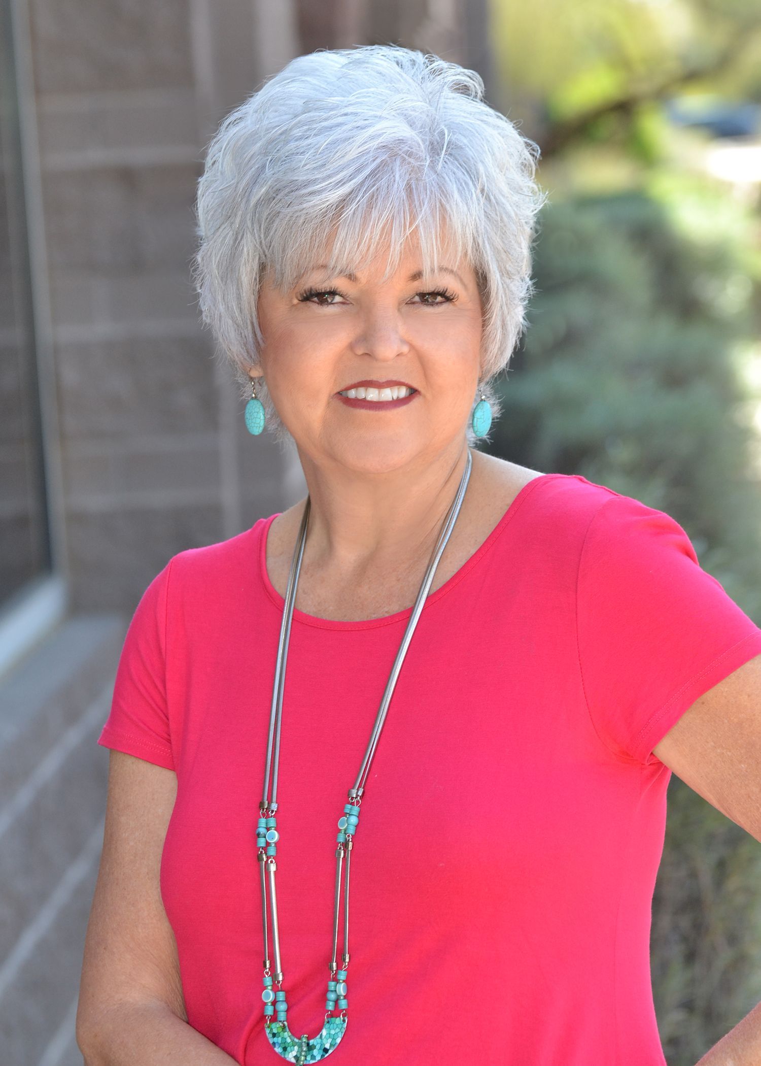 Portrait of a smiling woman in a blue top with silver hair and necklace against a black background