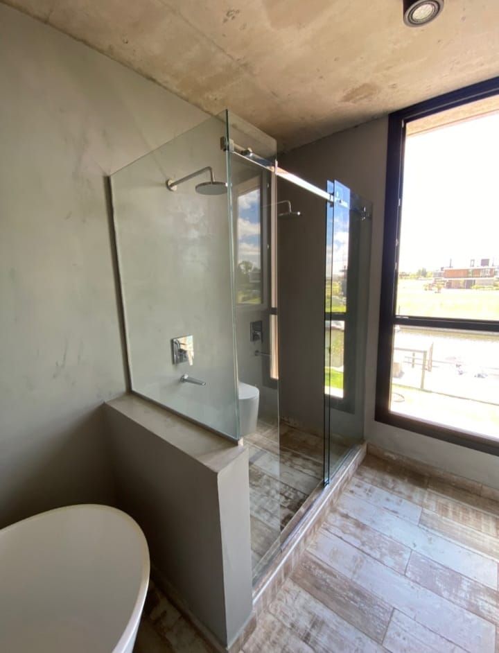 Baño moderno con mampara de ducha de cristal, paredes de hormigón y un gran ventanal.