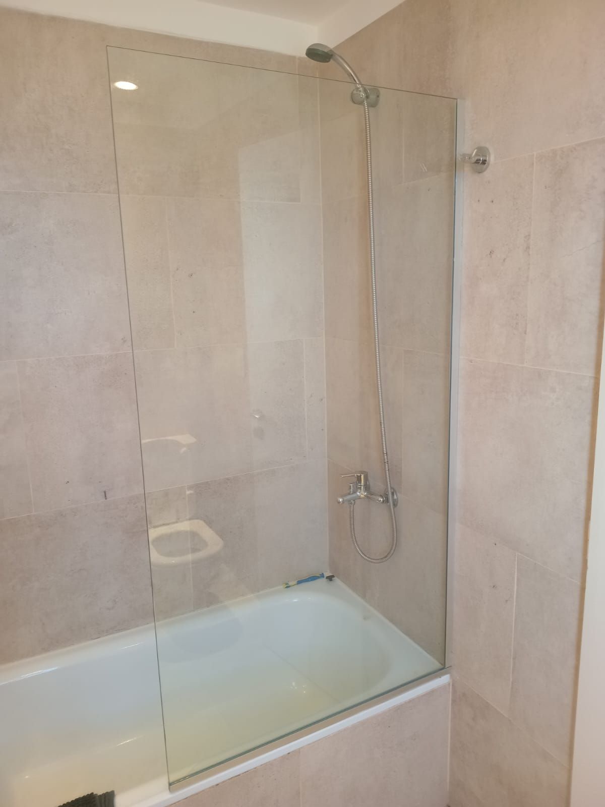 Baño con bañera blanca y mampara de cristal. Paredes de azulejos beige. Ducha y grifo plateados.