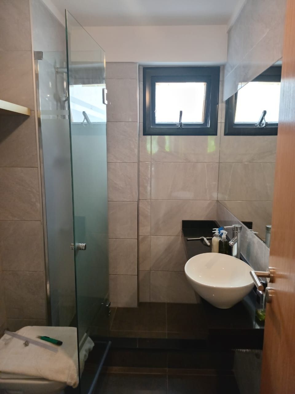 Baño con ducha, lavabo y ventana. Azulejos gris claro, lavabo blanco y mampara de cristal.