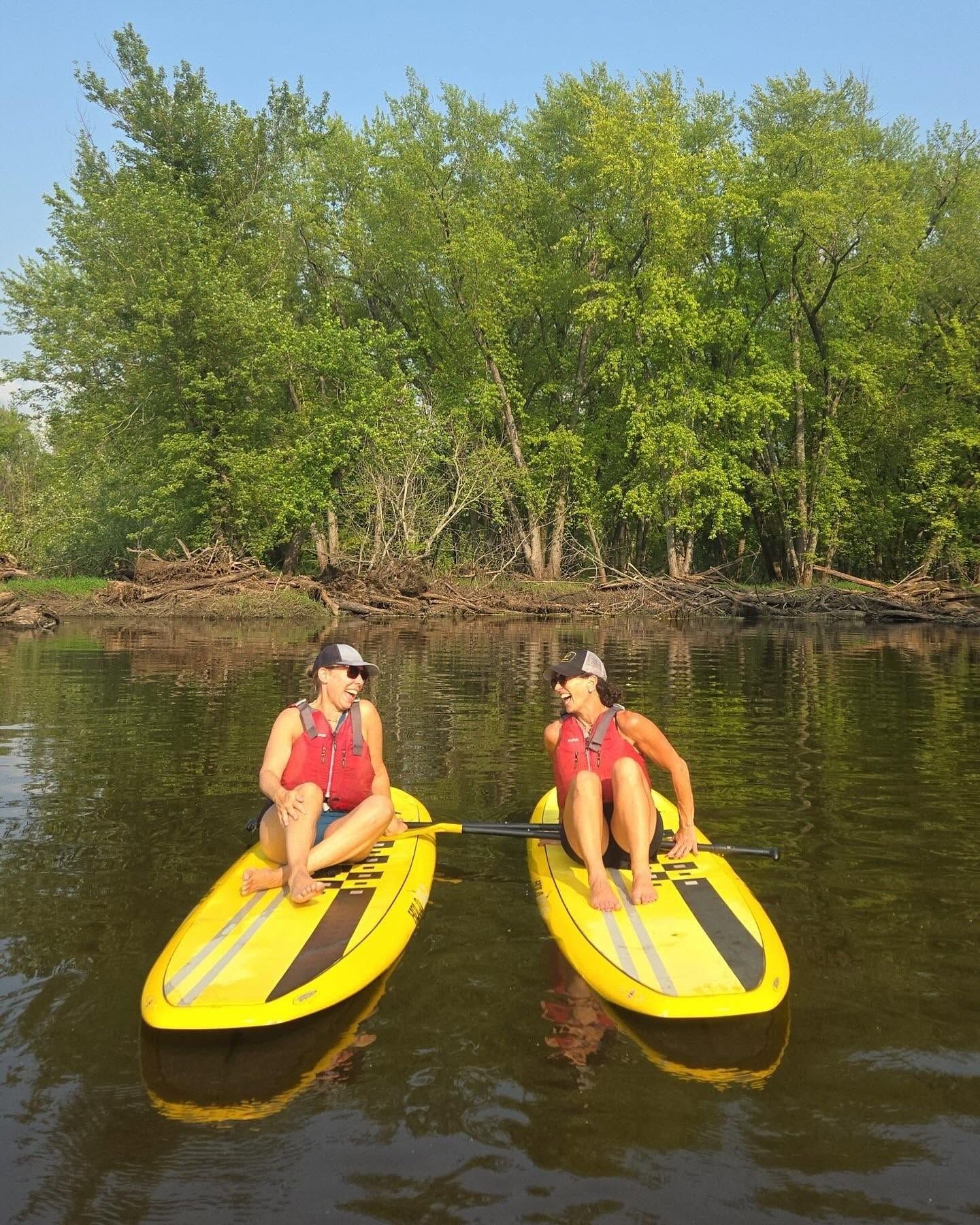 Two people on yellow kayaks on calm water beside a leafy riverbank.