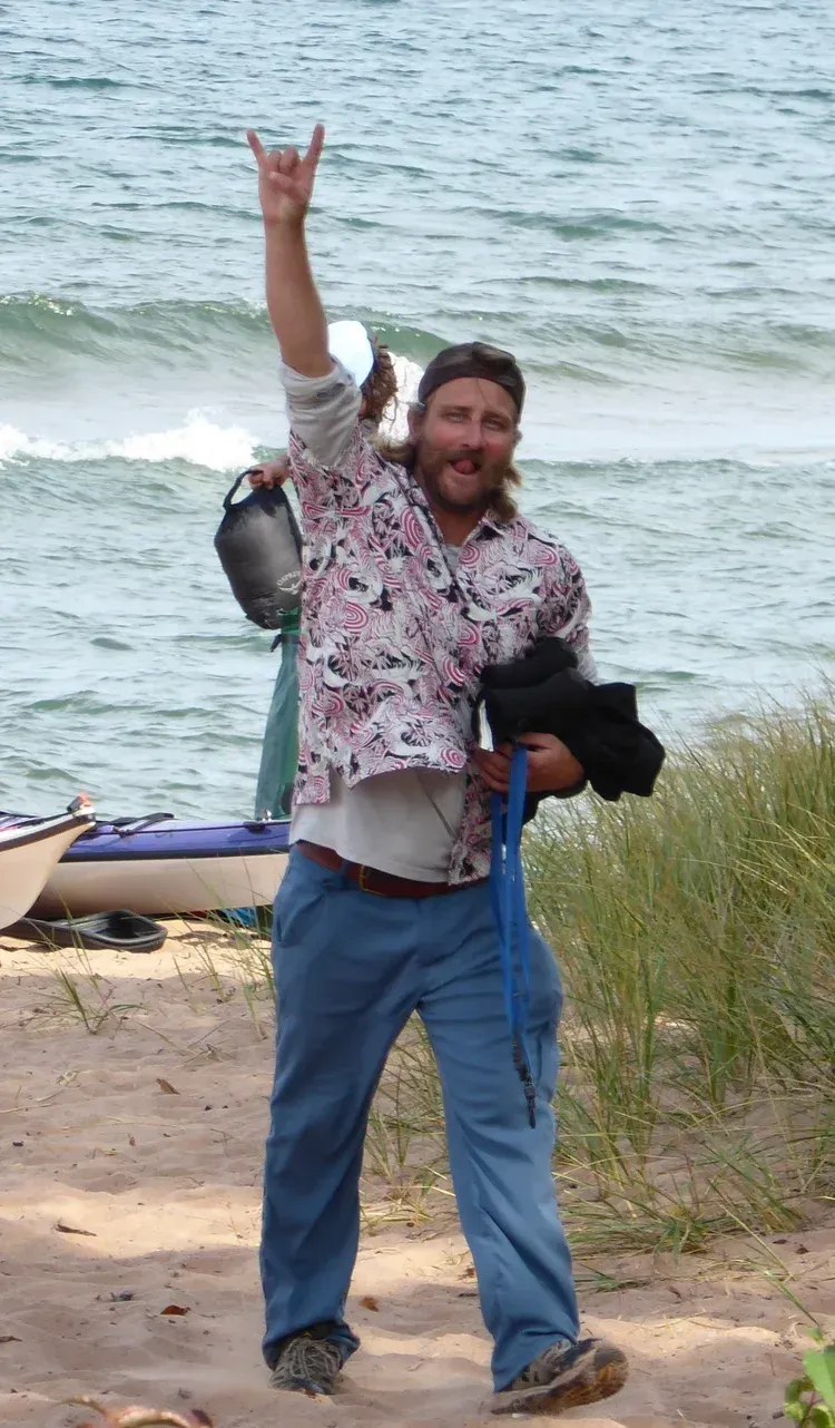 Smiling person in floral shirt and jeans raises a fish by a windy lakeshore