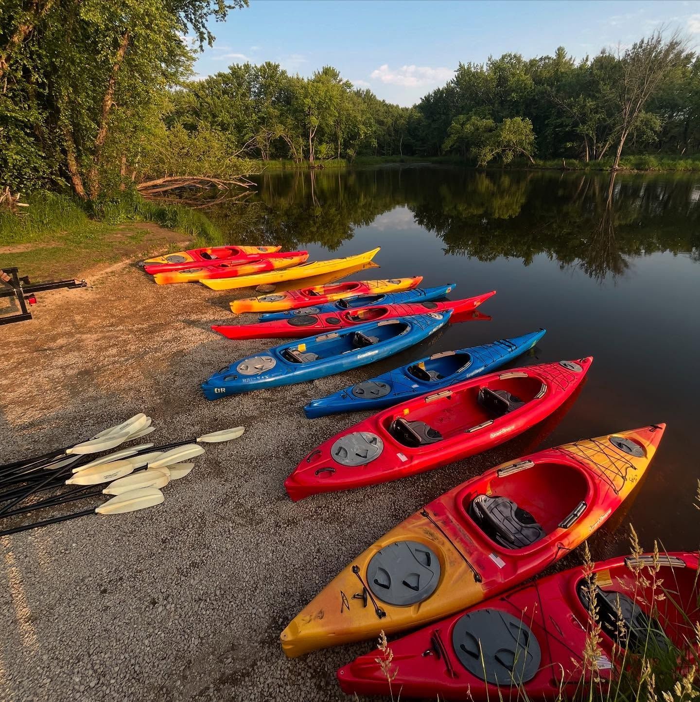 Colorful kayaks lined up on a pebbled shore beside a calm lake at sunrise.