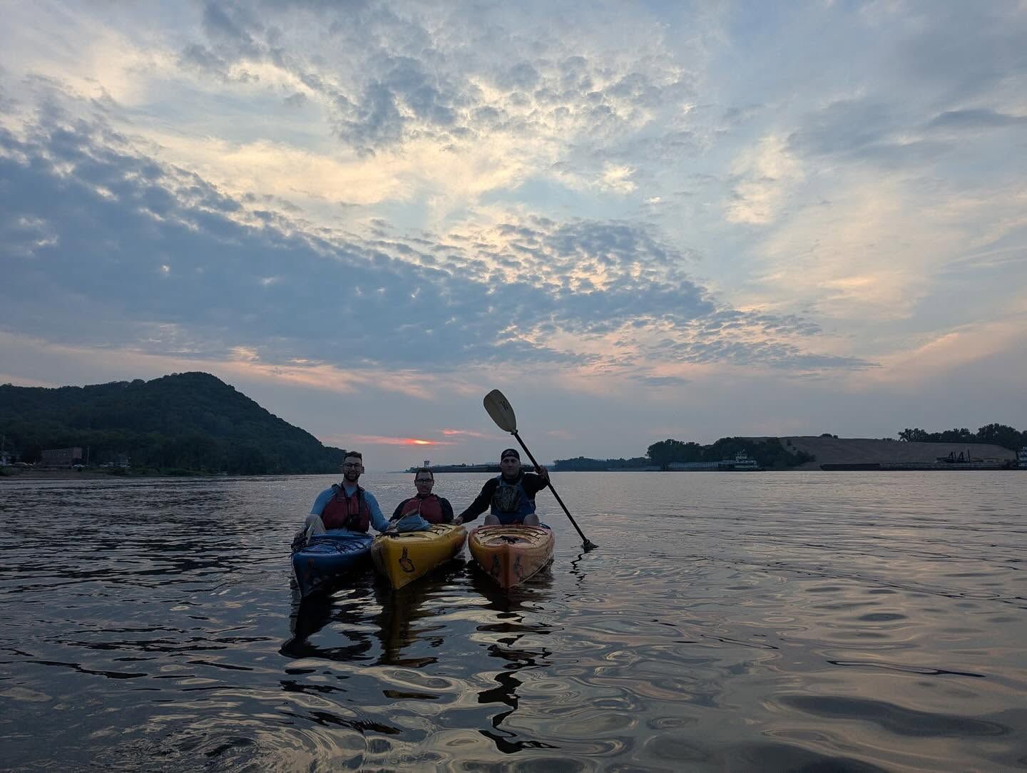 Two people paddle a canoe on calm water at sunset, with a distant shoreline and cloudy sky.