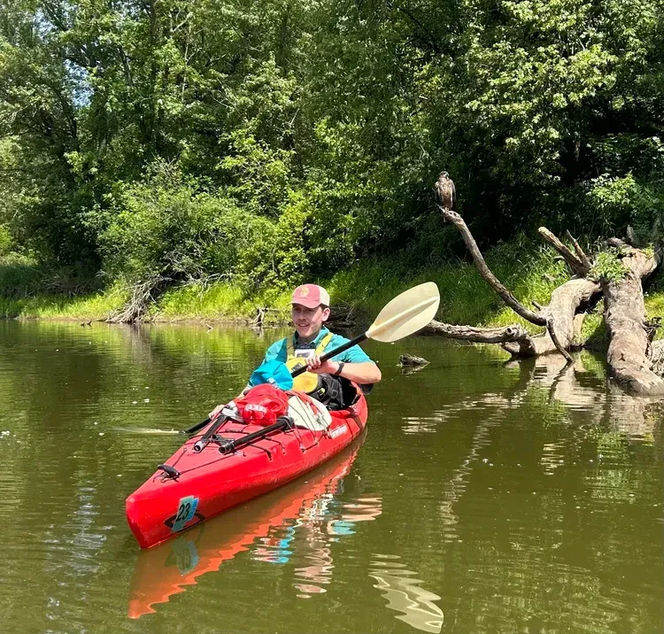 Person kayaking in a red kayak on a calm river, holding a paddle, with green trees along the bank