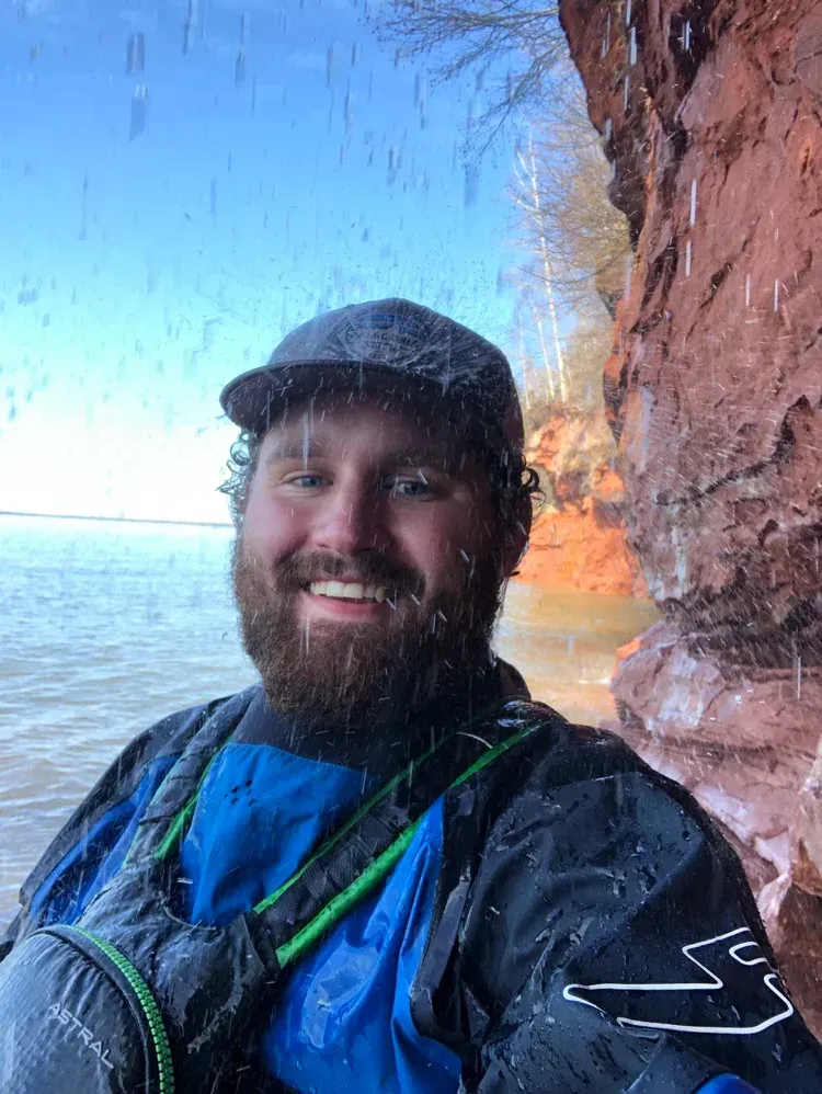 Smiling kayaker in blue jacket and black cap beside a red cliff with water and sky behind him