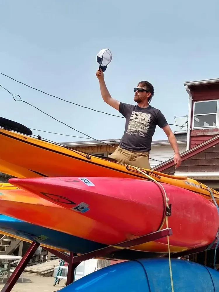 Man standing on a colorful kayak, raising a cap beside a beach with boats and a house in the background
