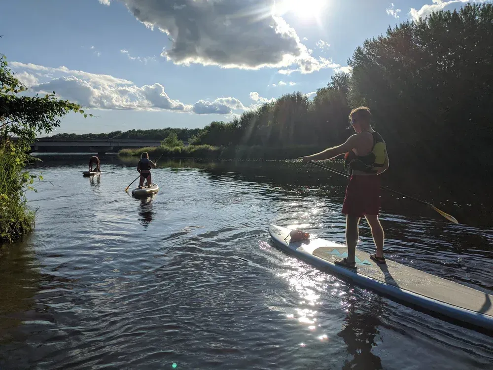 People paddleboarding on a calm river in bright sunlight, with trees and clouds along the shore.