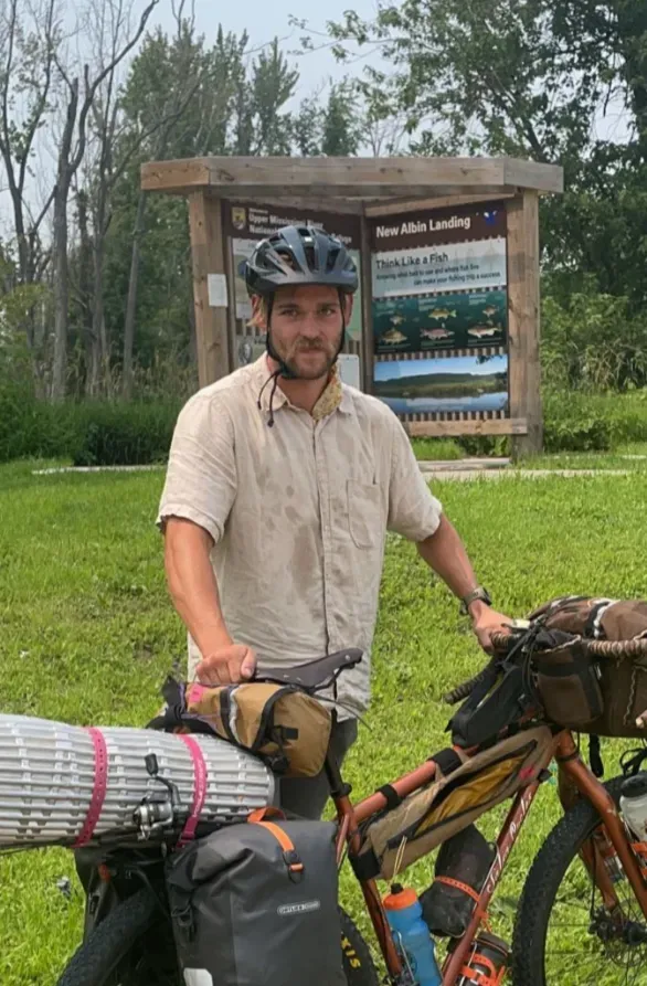 Bicycle traveler in a helmet stands by loaded bike near a wooden trail sign in a grassy park.