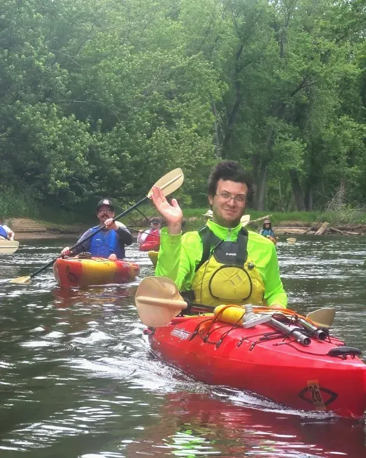 Kayakers paddling on a river; foreground paddler in bright green jacket on a red kayak, wooded banks behind.