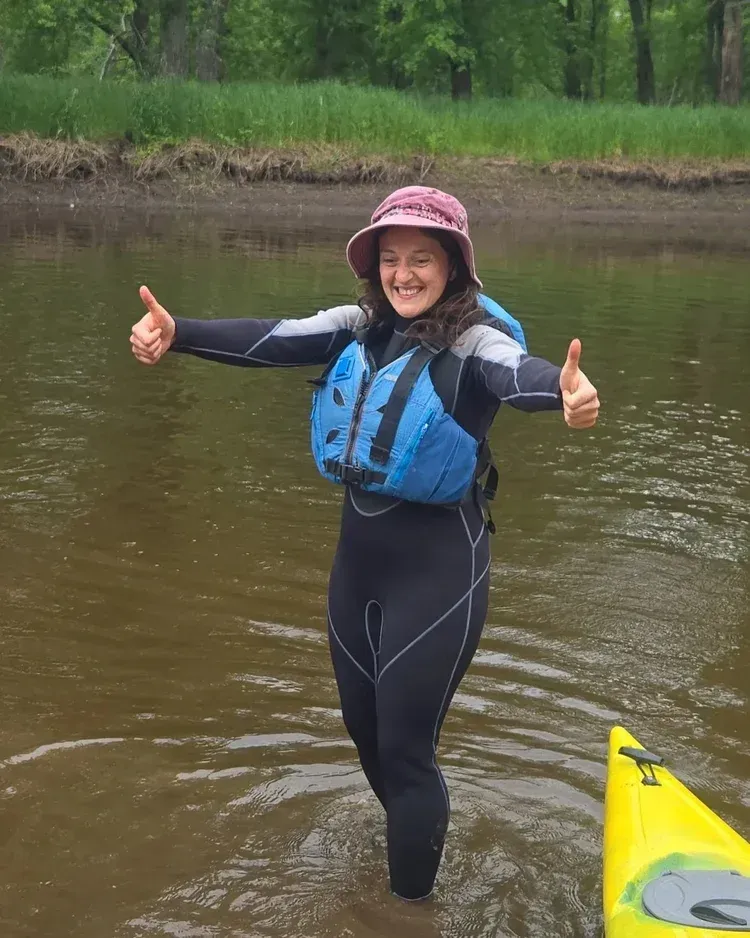 Smiling person in a blue wetsuit and pink helmet gives thumbs up while standing in a river by a kayak