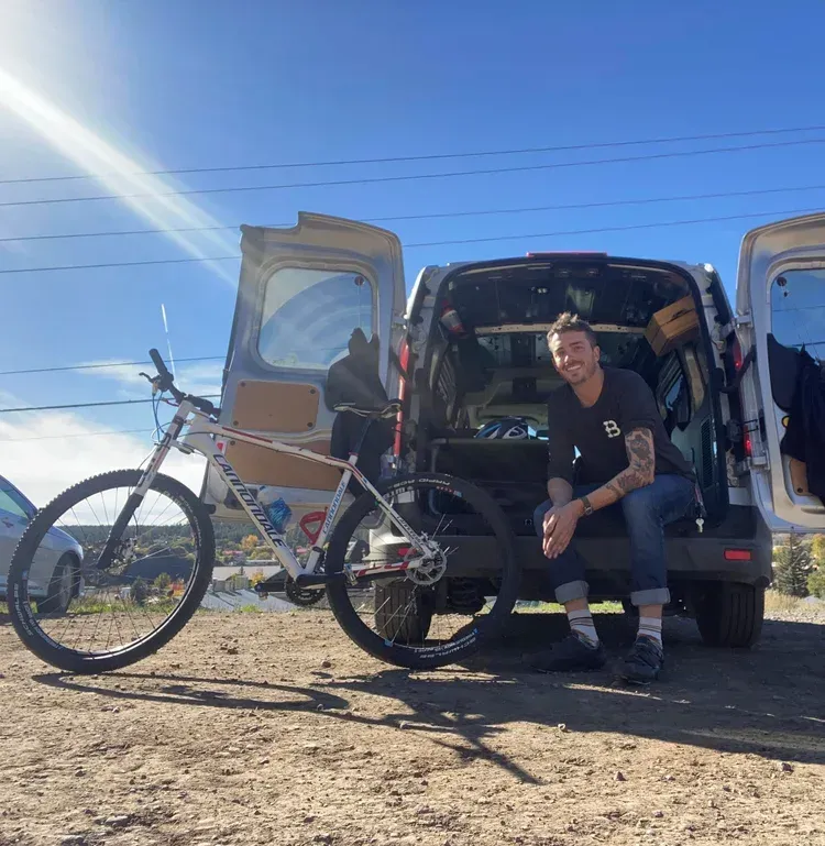 Man sitting beside a van with a bicycle, bright blue sky and open doors in a dusty outdoor setting