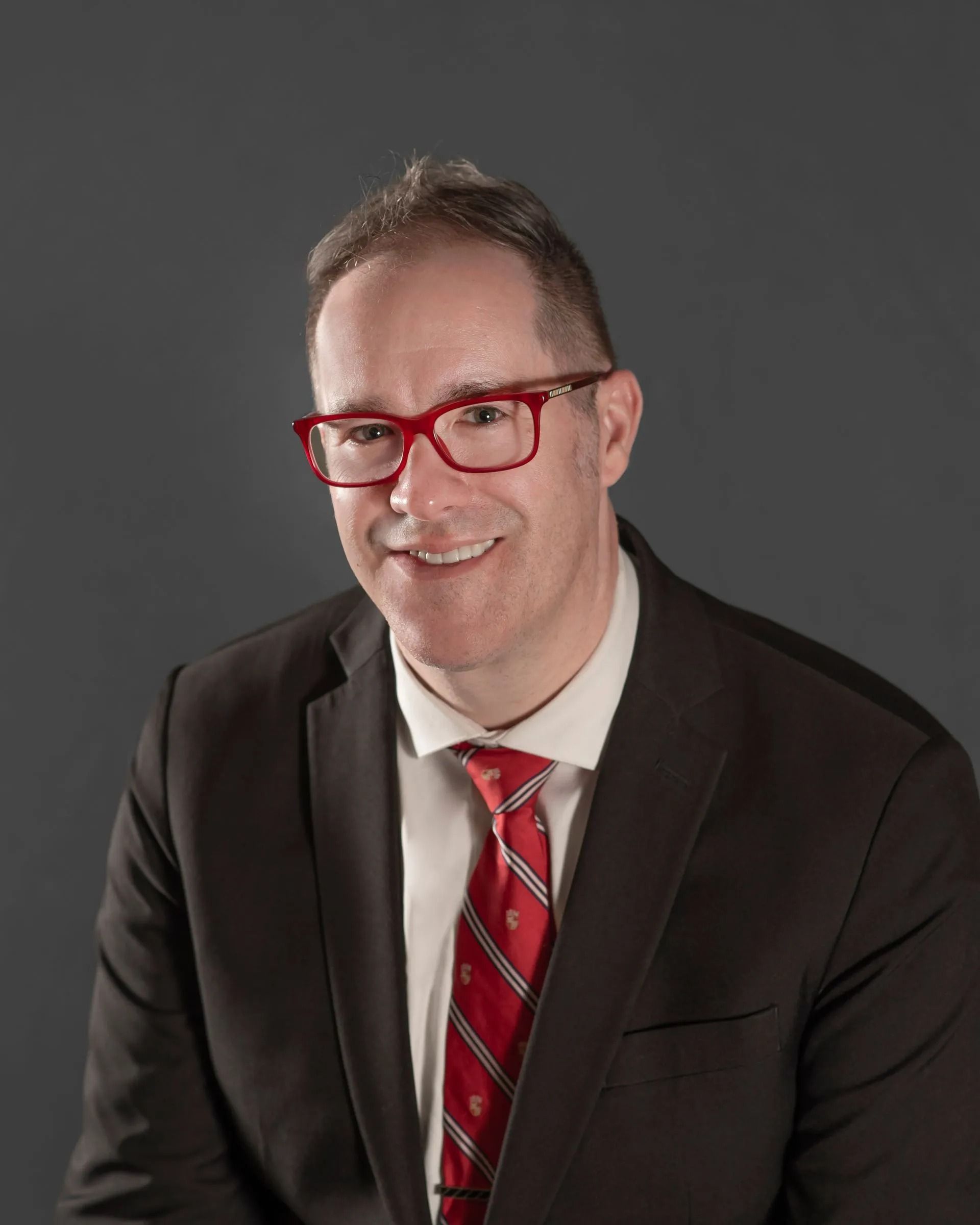 Man in a dark suit, white shirt, and red striped tie, wearing red glasses, smiling against a gray backdrop