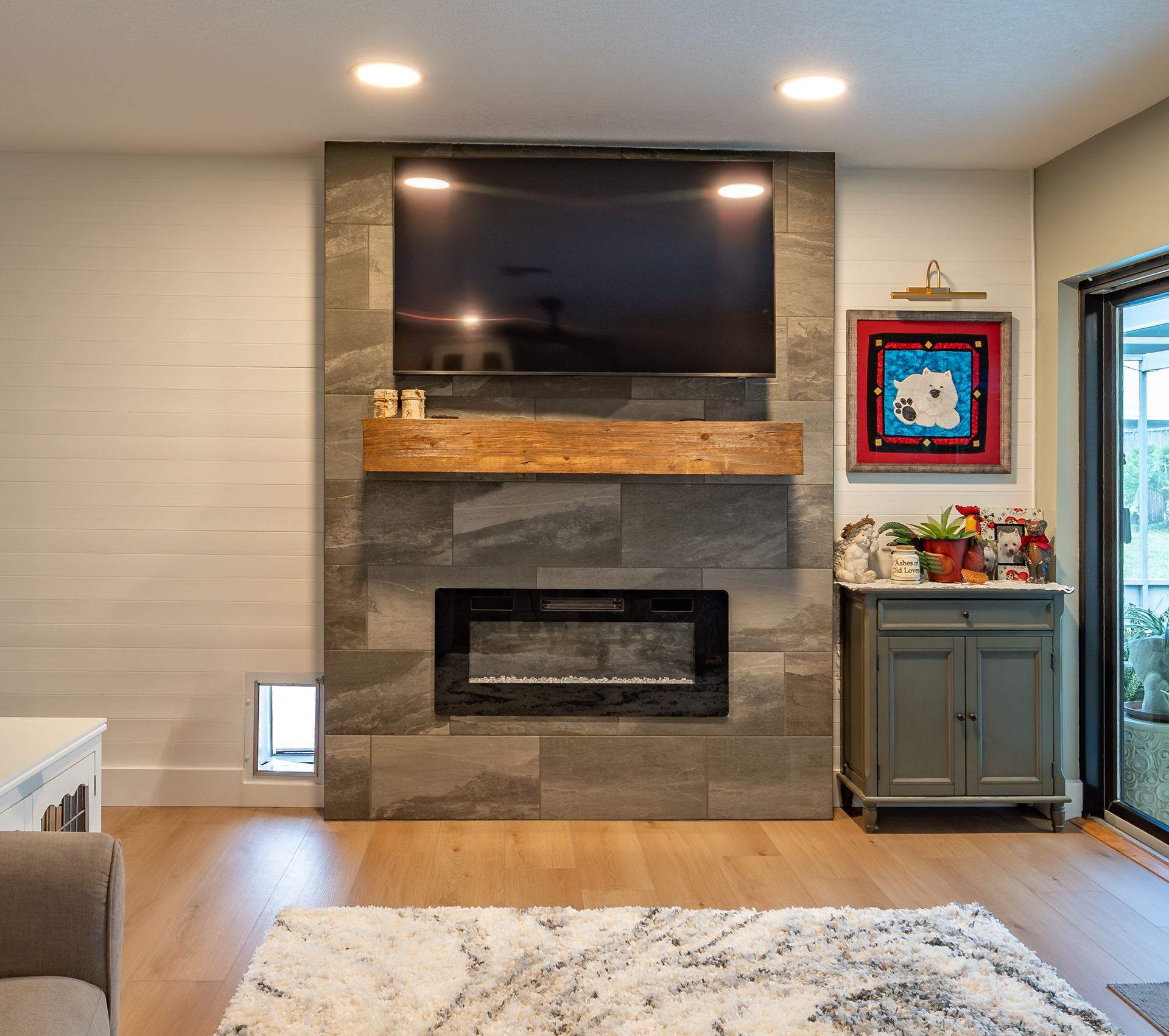 Living room with fireplace and TV on a gray stone wall; wooden mantel; dog door; rug.