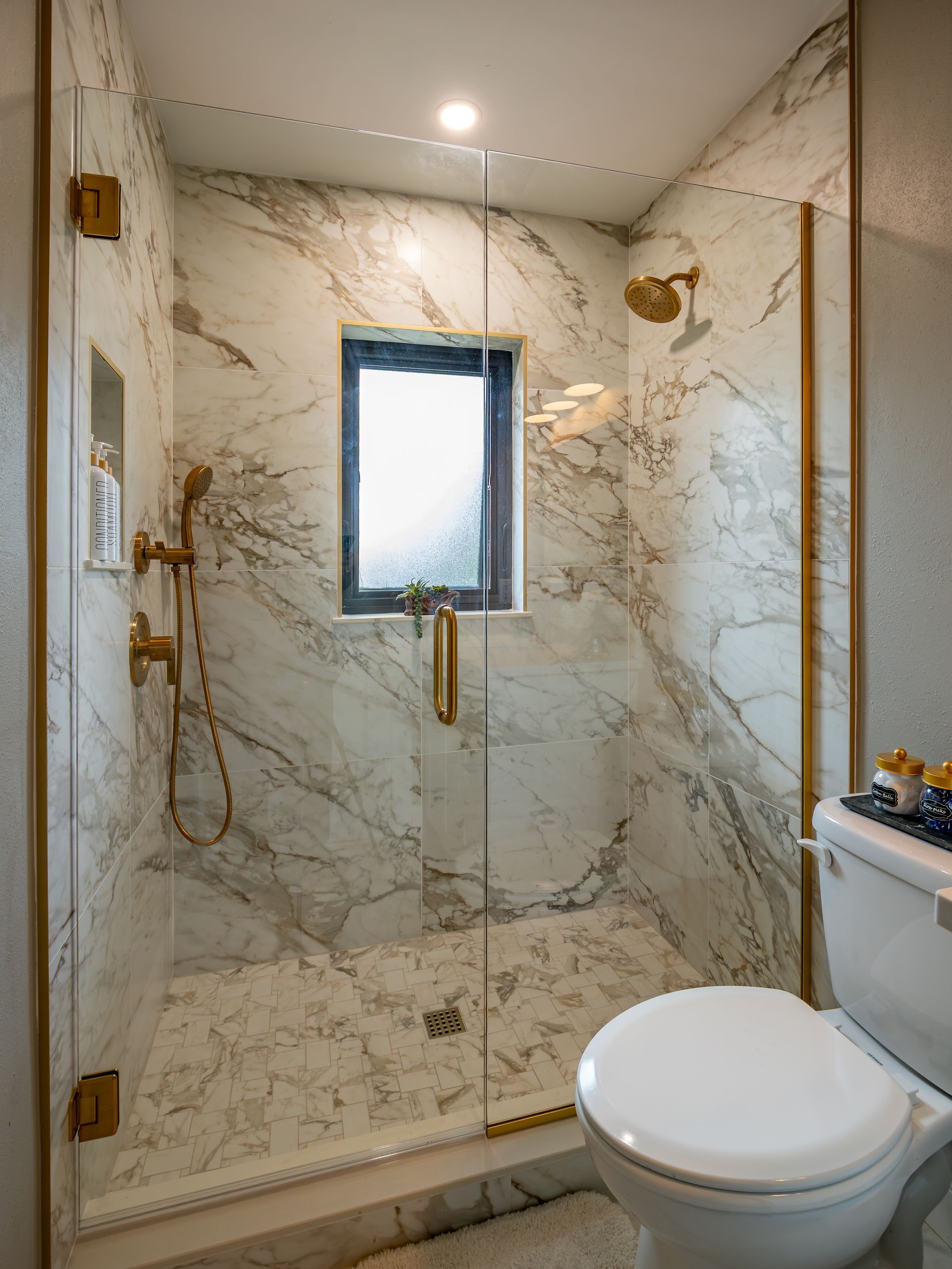 Marble-tiled shower with gold fixtures, glass doors, and a small window. A white toilet sits to the side.