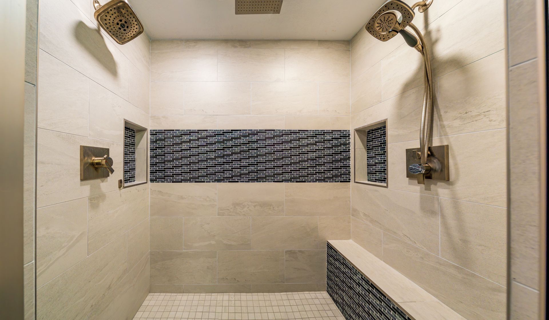 Shower with two showerheads, built-in bench, and inset blue tiled band against cream-colored tile.