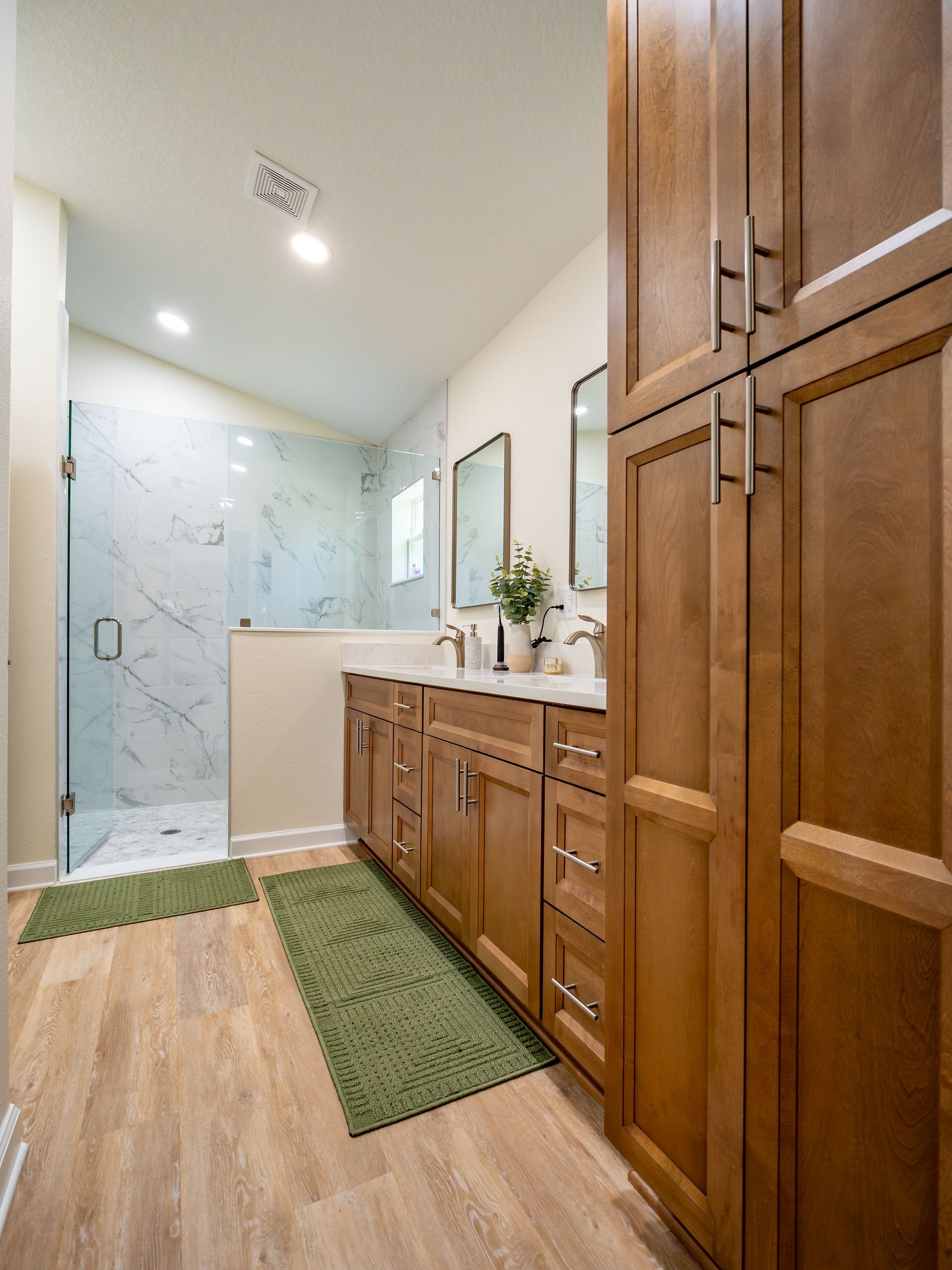 Bathroom with wooden cabinets, double vanity, glass shower, and wooden floor.