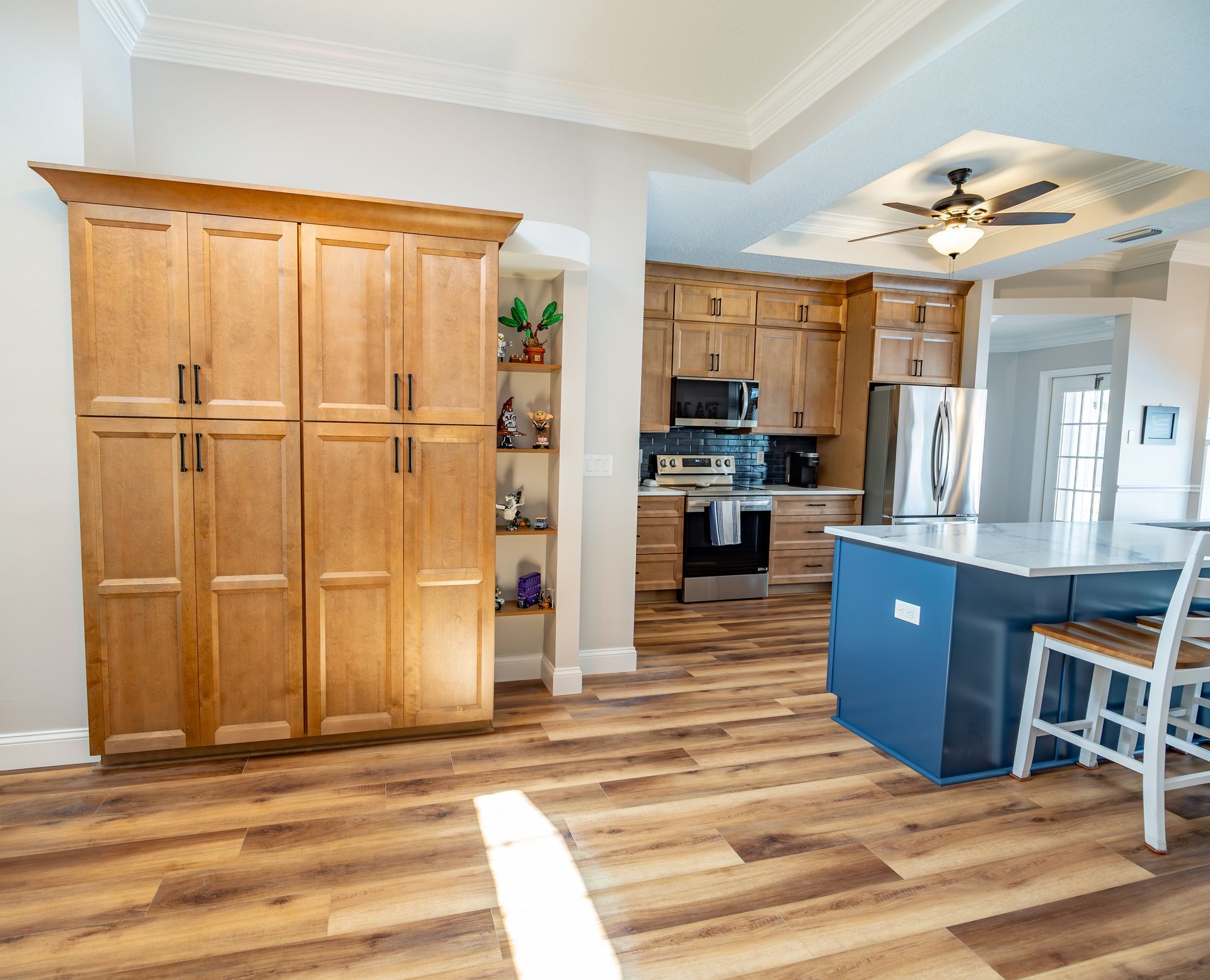 Wooden pantry cabinet in a kitchen, adjacent open shelving. Kitchen has blue island, wood floors, beige walls.