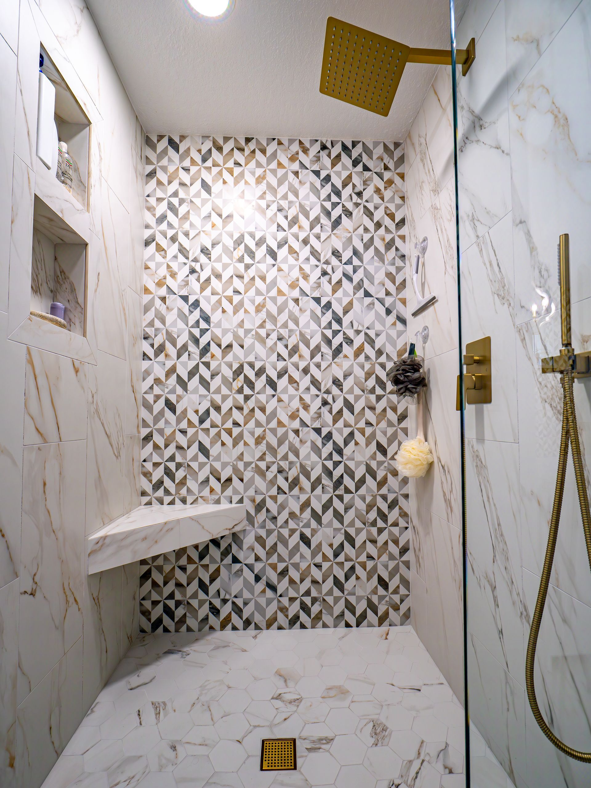 Modern shower with gold fixtures, marble-like tiles, and accent wall with geometric pattern.
