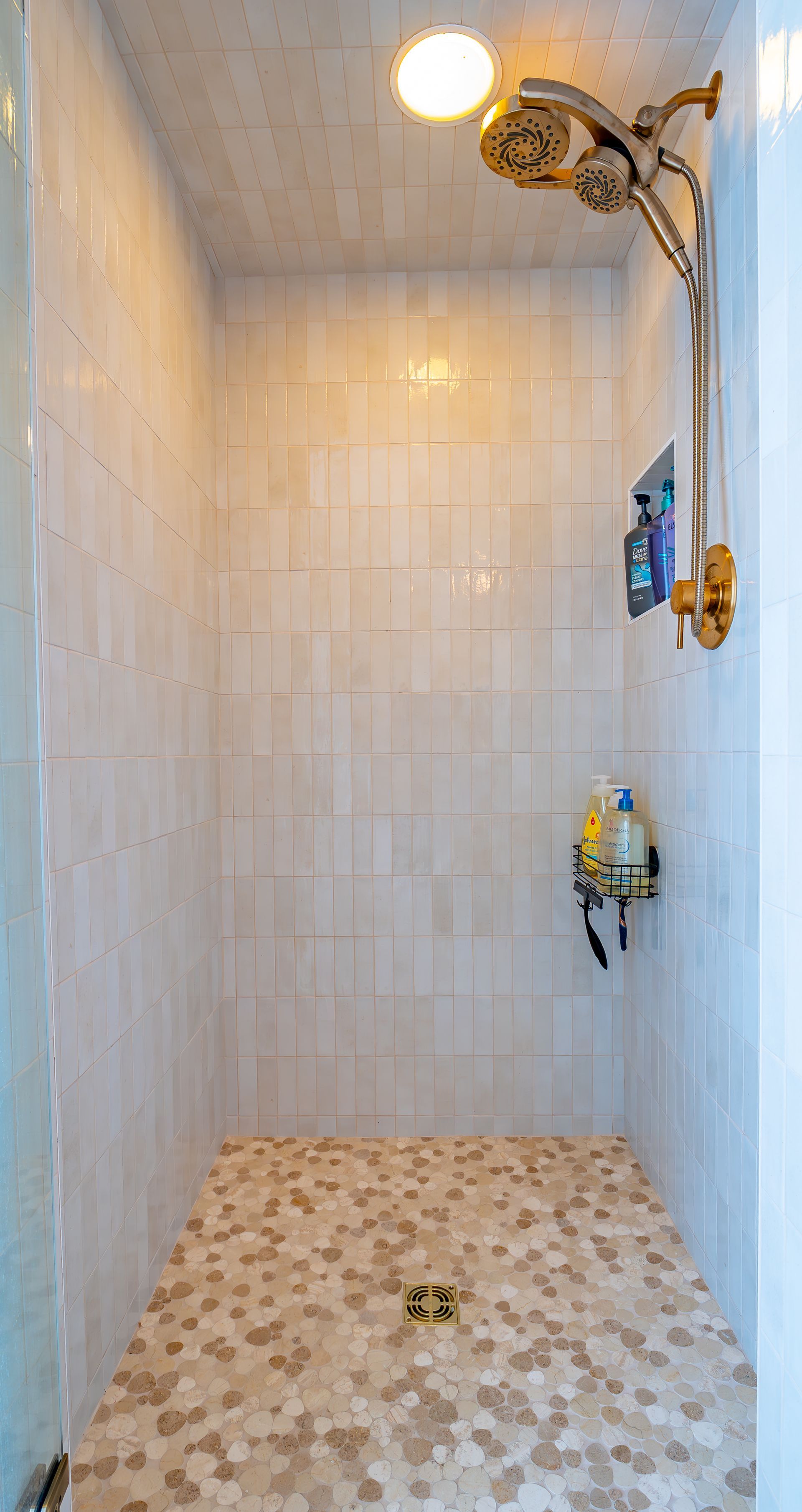 Shower stall with stone floor, beige tiled walls, and gold shower fixtures.