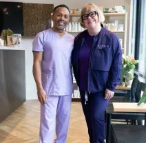 Two people standing together in a bright café, wearing purple outfits and smiling at the camera.
