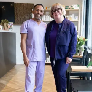 Two people standing together in a bright salon, one in lavender scrubs and one in a navy jacket.