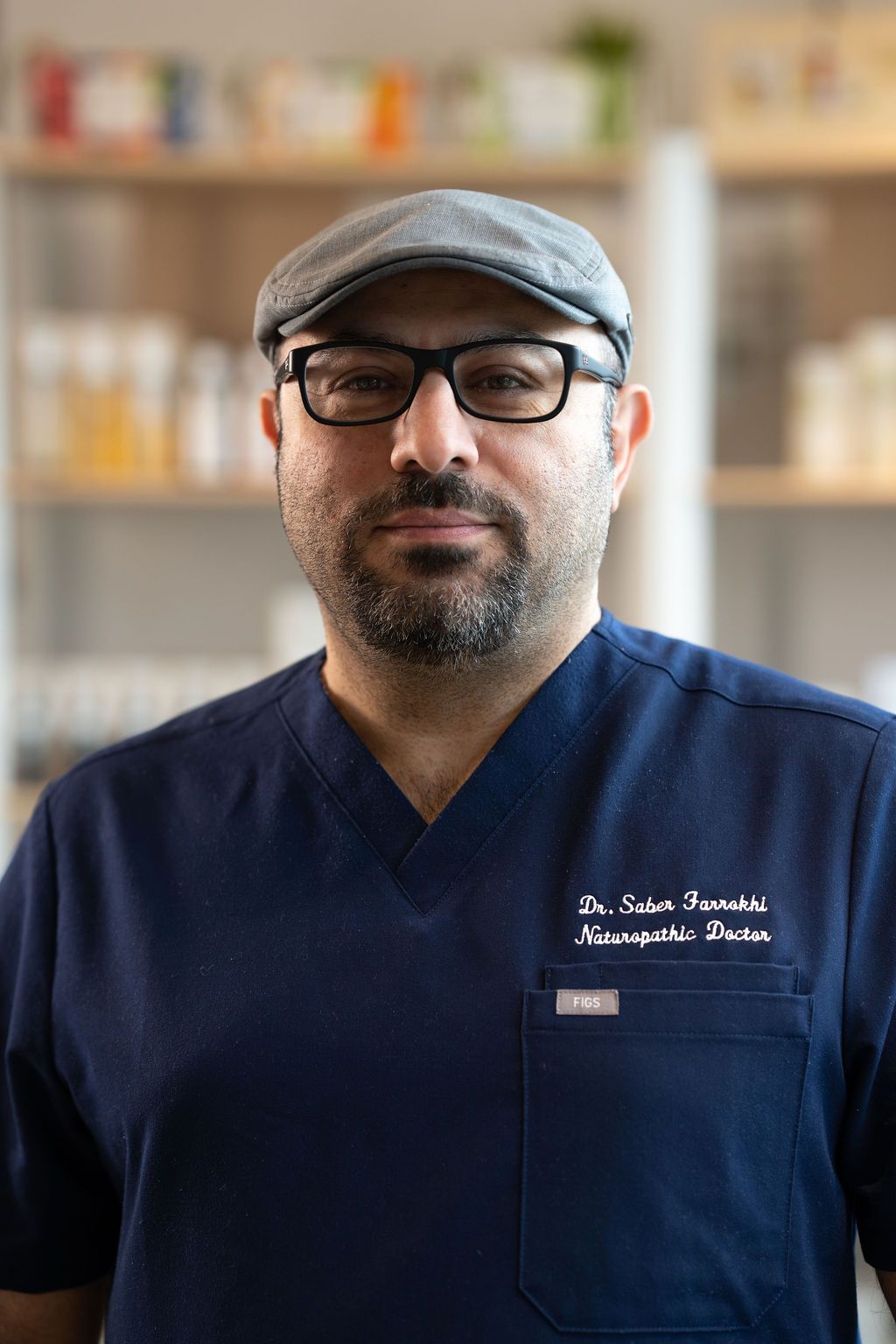 Portrait of a man in navy scrubs and cap, wearing glasses, standing in a clinic with blurred shelves behind him