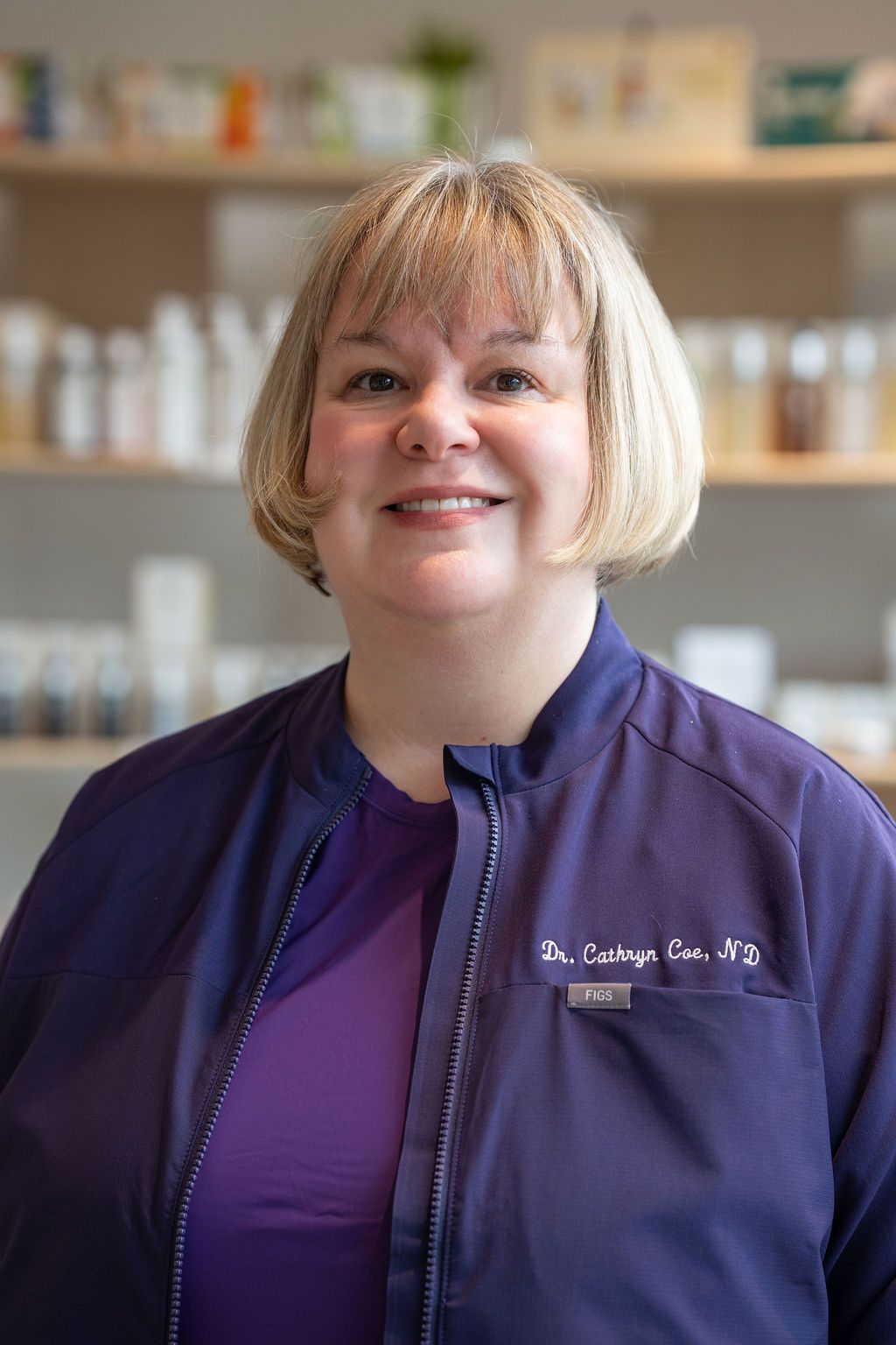 Smiling person in a purple top and navy vest standing in front of pharmacy shelves