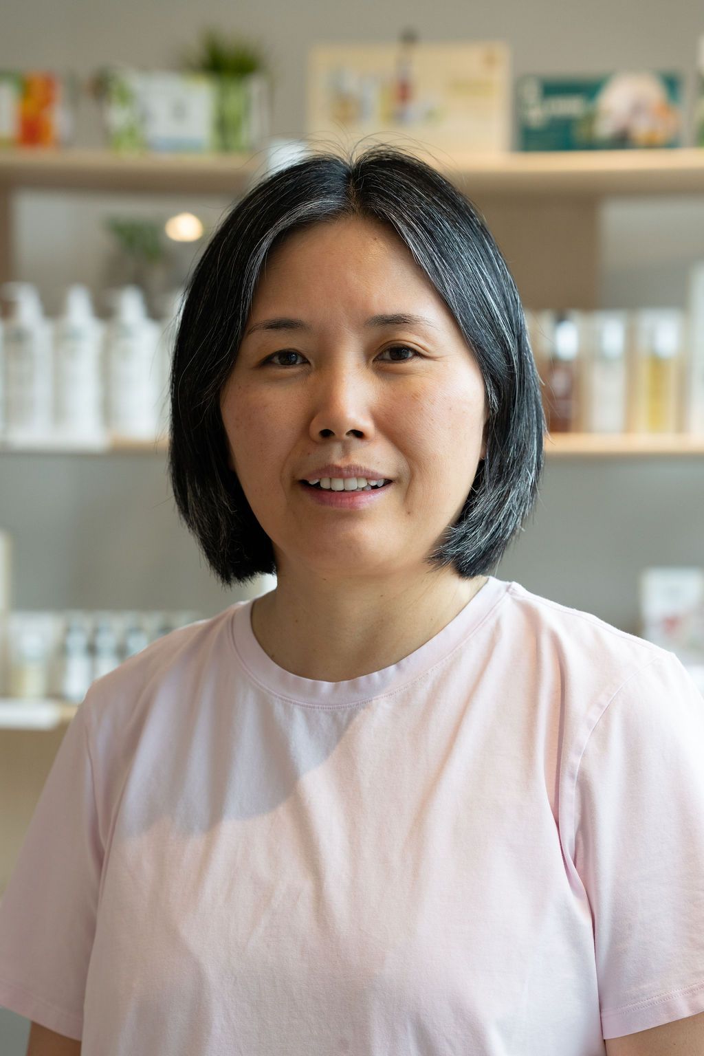Portrait of a person with short black hair in a light pink shirt, standing in front of shelves of bottles