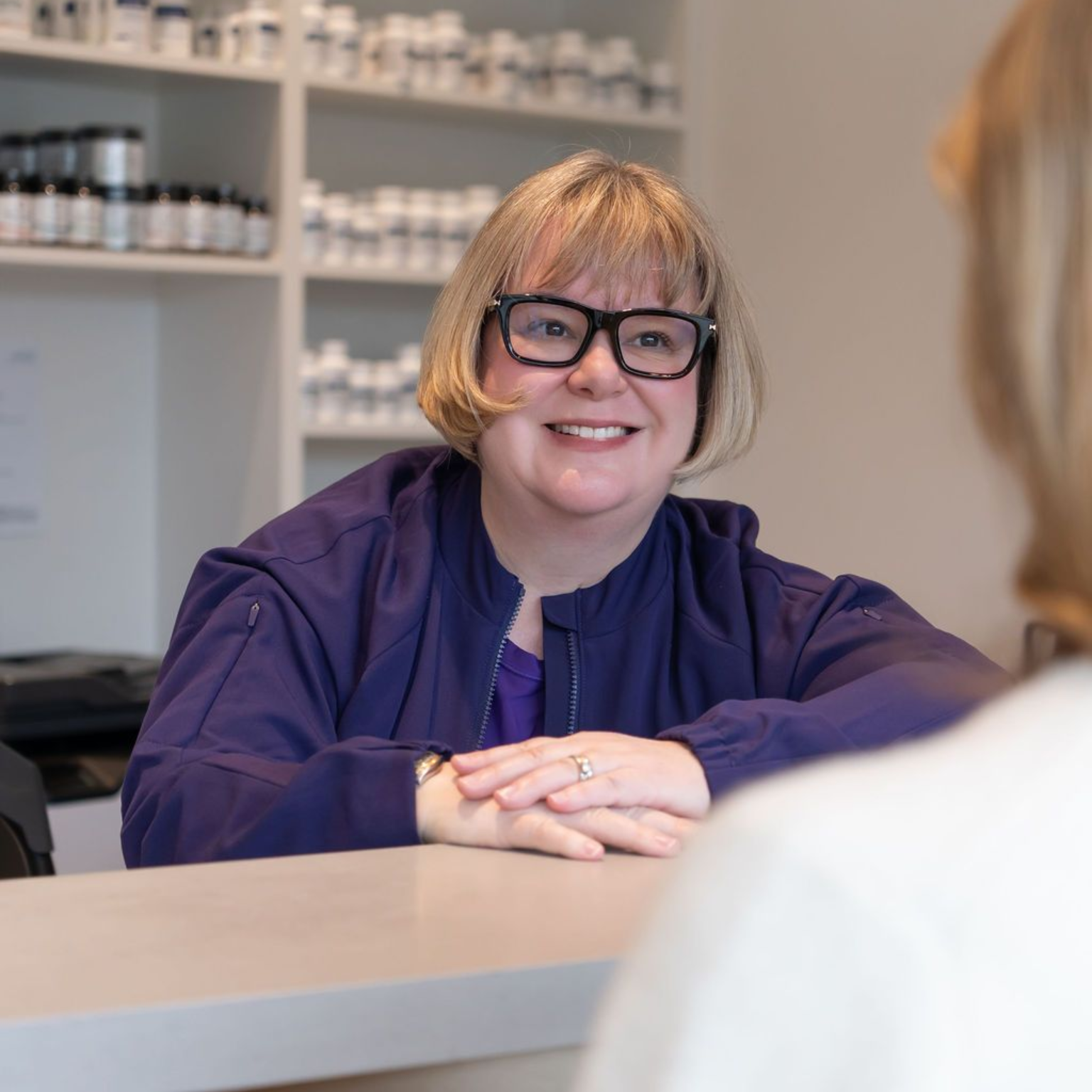 Smiling receptionist in purple top behind a counter, speaking with a customer in a bright office