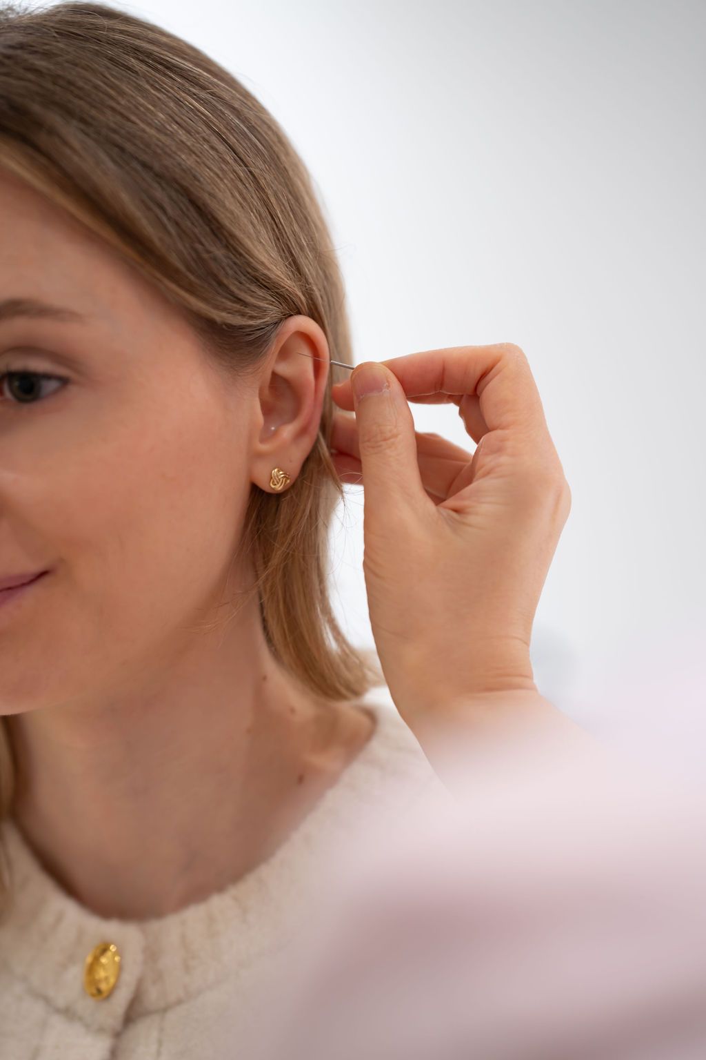 Woman adjusting a gold hoop earring, wearing a white top against a light background