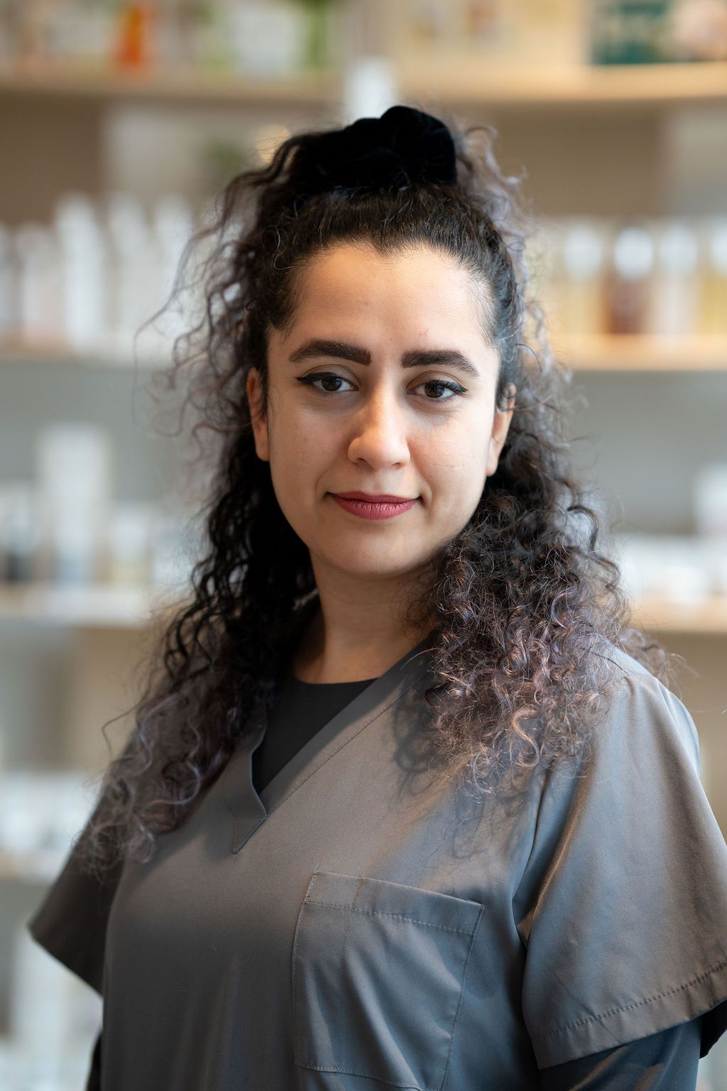 Portrait of a person with curly hair in a gray top, standing in a softly blurred indoor setting.