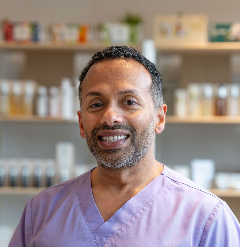 Smiling person in purple scrubs standing in a pharmacy with shelves of medicine behind them