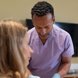 Two people in a clinic, one in purple scrubs leaning over a desk and speaking to the other.