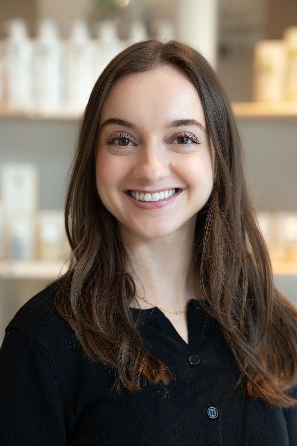 Smiling woman in a black top standing in front of softly blurred shelves in a bright salon-like setting