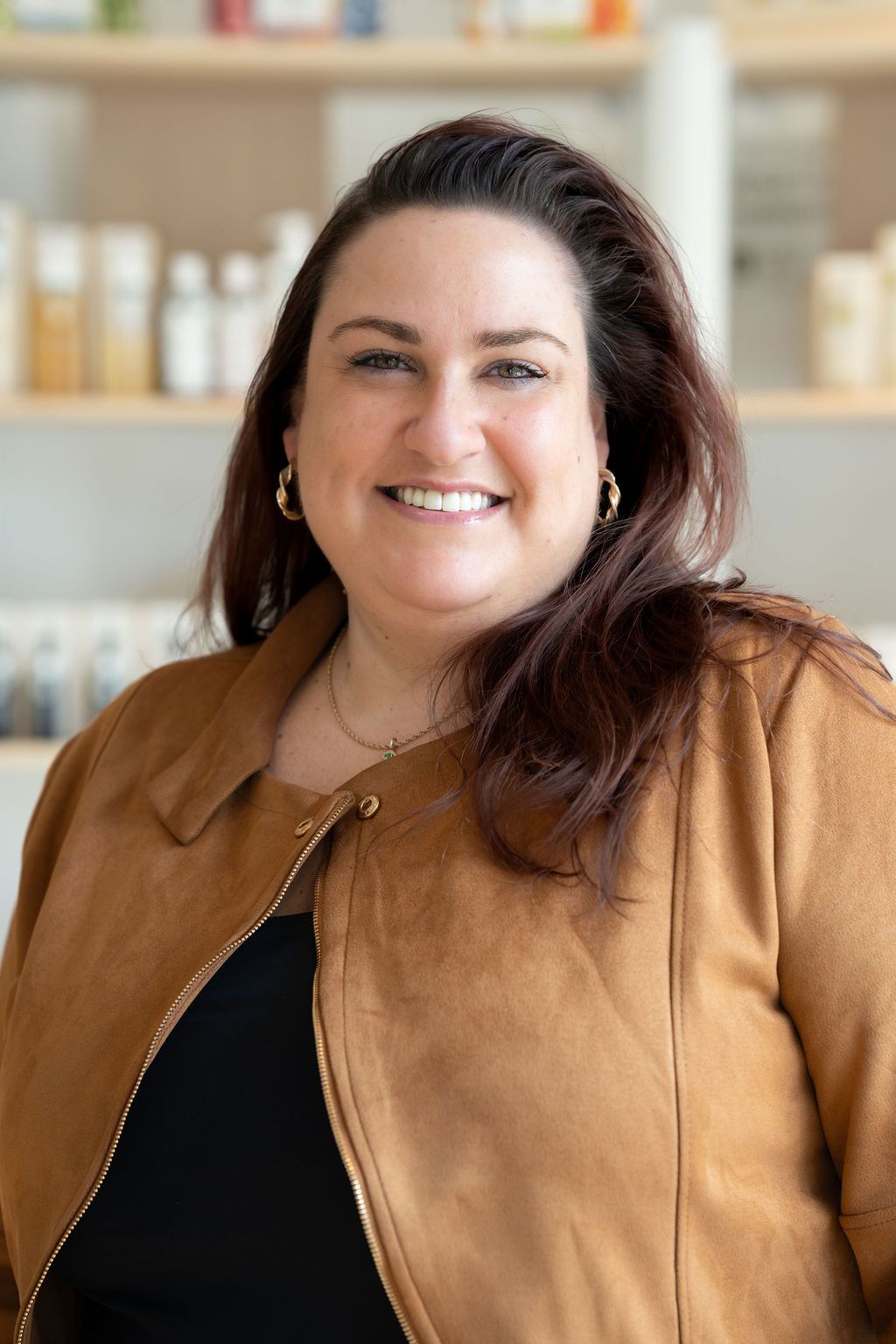 Smiling woman in a tan jacket standing indoors, with shelves blurred in the background.
