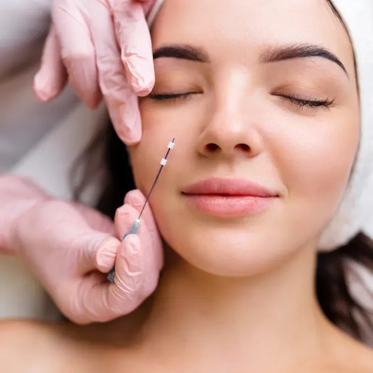 Gloved hands hold a small syringe near a woman’s cheek during a cosmetic facial treatment
