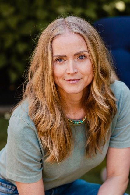 Portrait of a smiling woman with long blonde hair in a green shirt, sitting outdoors against a blurred background