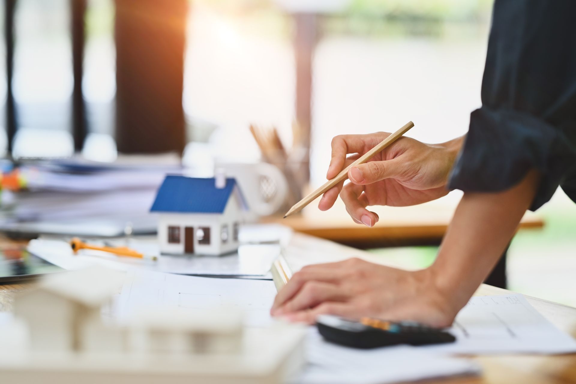 An architect works at a desk, sketching on blueprints next to a small house model and drafting tools.
