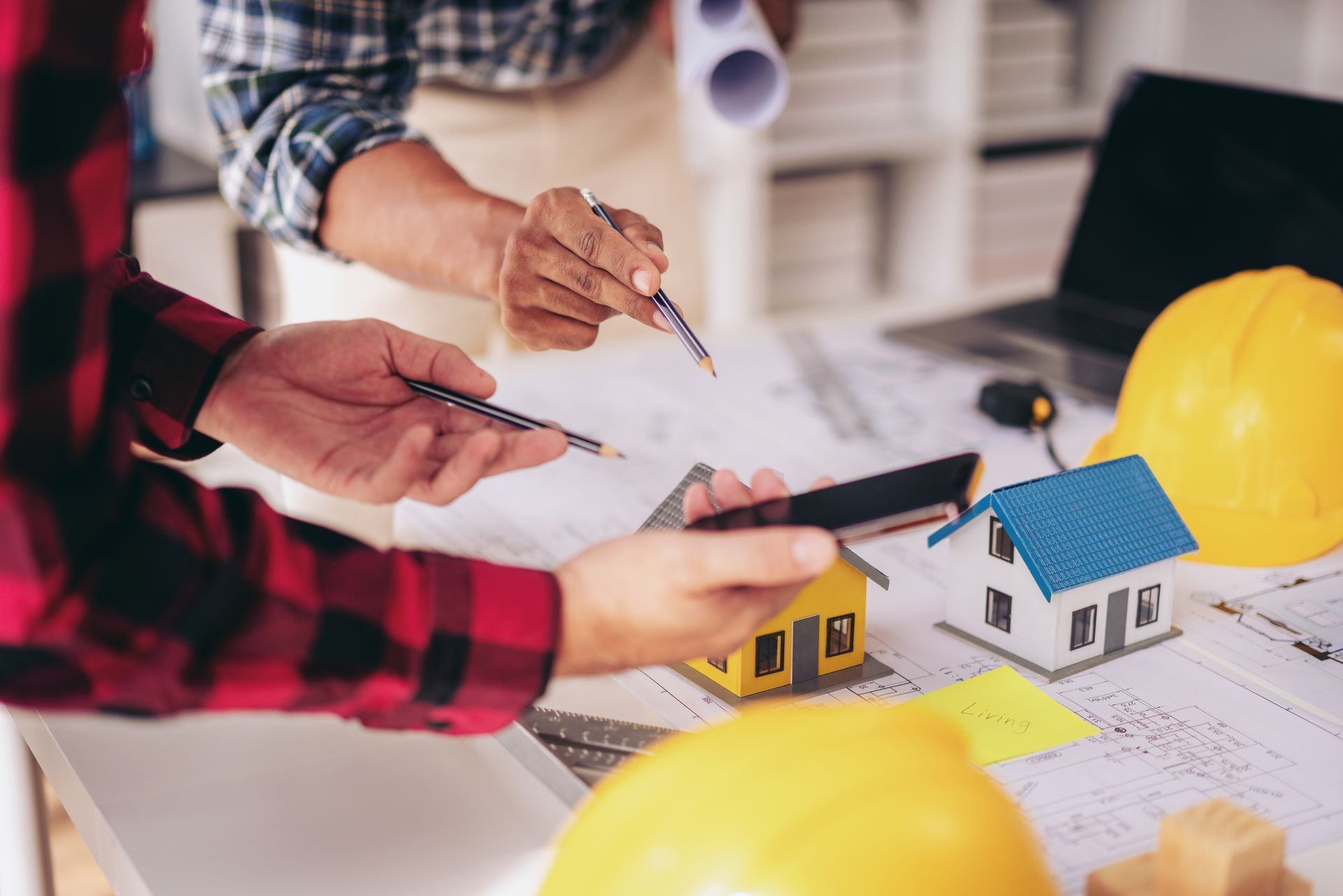 Two people collaborate on architectural plans, holding a model house and pointing to blueprints beside a yellow hard hat.
