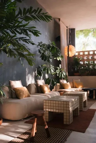 Cozy outdoor lounge with beige couches, tiled tables, and lush greenery; neutral tones, natural light.