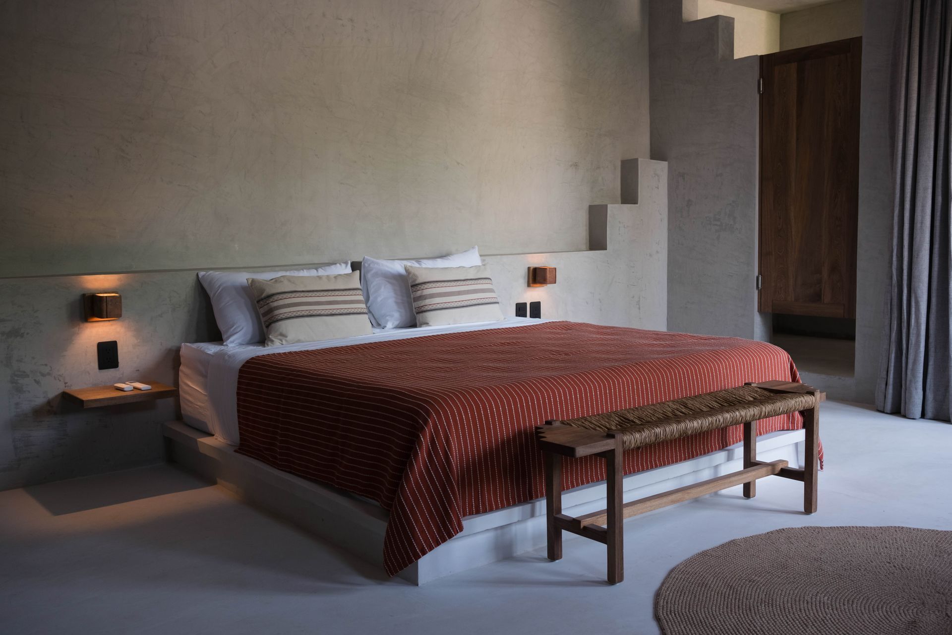 Bedroom with a large bed, orange patterned blanket, wooden bench, and gray walls.