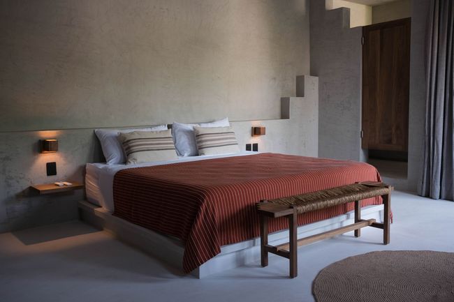 Bedroom with a large bed, orange patterned blanket, wooden bench, and gray walls.