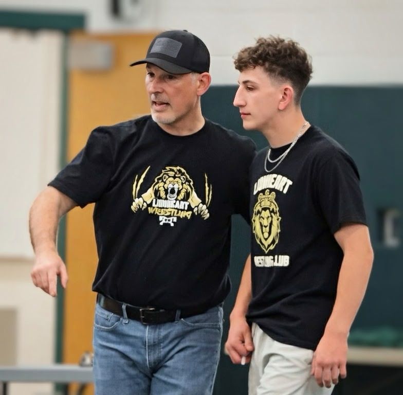 Wrestling coaches instructing a group of children on a wrestling mat in a gym.